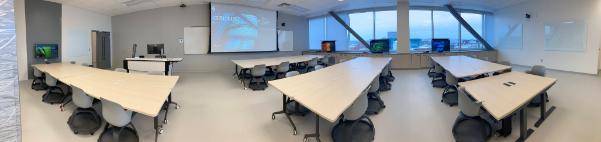 A photo of a classroom with tables with moveable chairs around them and a large pull-down projector screen at the front.